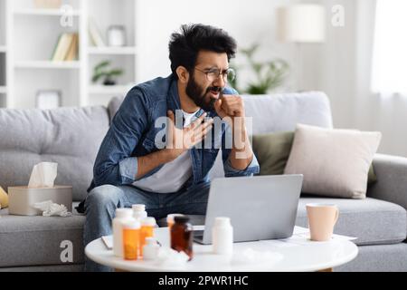 Homme indien malade qui tousse dans le poing tout en étant assis sur le canapé à la maison Banque D'Images