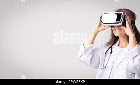 Médecin senior femme portant des lunettes de réalité virtuelle, debout sur fond gris studio, panorama avec espace de copie Banque D'Images