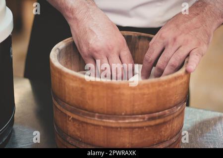 Gros plan des mains d'un chef sushi méconnaissable cuisant du riz dans un bassin ou un seau en bois asiatique traditionnel Banque D'Images