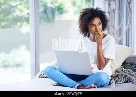 Grignoter des en-cas sains tout en naviguant sur le Web. une jeune femme mangeant une pomme tout en utilisant son ordinateur portable à la maison. Banque D'Images