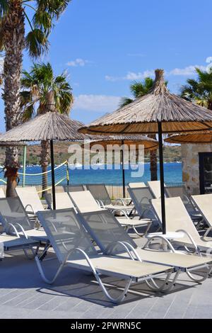 Transats vides, palmiers et parasols bambu au bord de la piscine avec la mer à l'arrière-plan dans une journée ensoleillée d'été Banque D'Images