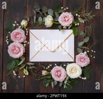 Enveloppe entre roses rose et crème sur bois brun vue de dessus, maquette de mariage. Scène romantique avec enveloppe et fleurs pastel. Saint-Valentin, S. Banque D'Images