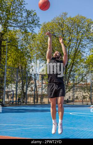 Athlétique homme libanais dans les vêtements de sport jouant au basket-ball, lance avec succès le ballon dans l'anneau de panier. Jeune homme sur le terrain d'été de la ville urbaine. Programme de fitness sur le terrain de sport. Motivation. À l'extérieur Banque D'Images