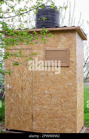 Une cabine de douche faite de panneaux OSB, avec un grand baril pour chauffer l'eau du soleil, dans la cour de la maison au printemps. Création d'un spectacle d'été Banque D'Images