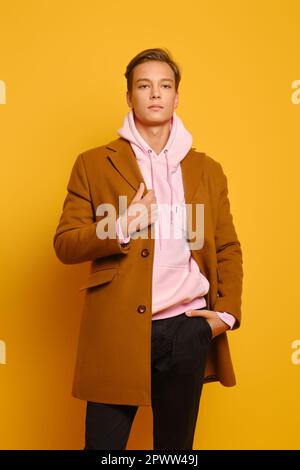 Beau homme élégant en manteau brun et pull à capuche rose posé sur fond jaune studio Banque D'Images