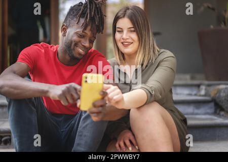 Deux amis divers, un Africain et une femme de taille plus, assis sur des escaliers extérieurs, regardant un contenu de smartphone lors d'une journée ensoleillée d'été. Banque D'Images