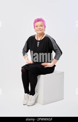 Une vieille femme souriante avec des cheveux roses courts en robe tricotée longue se trouve directement sur une boîte blanche dans le studio Banque D'Images