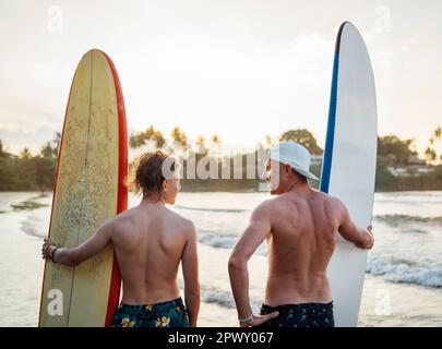 Père avec un fils adolescent debout avec des planches de surf sur la plage de sable de l'océan avec des palmiers sur fond éclairé par le soleil de coucher de soleil.Ils sourient et h Banque D'Images