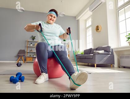 Graisse obèse femme faisant des exercices d'étirement avec bande de caoutchouc assis sur la balle de forme à la maison. Banque D'Images