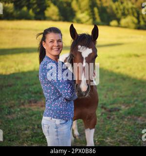 Femme en Jean et chemise debout à côté de brun et blanc pinto cheval foal, vert prairie derrière elle, photo de gros plan Banque D'Images