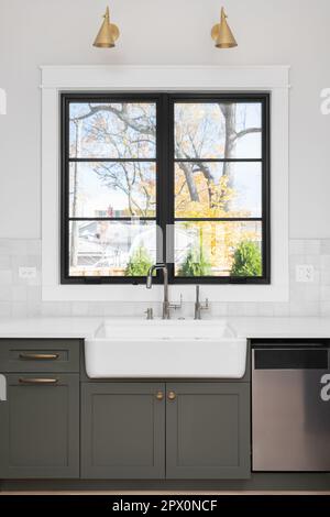 Un détail d'évier photographié avec un évier de tablier, des armoires vertes, un cadre de fenêtre noir, un dosseret carrelé, et des appliques dorées au-dessus de la fenêtre. Banque D'Images