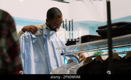 Client afro-américain qui se charge de vêtements sur cintres, qui regarde la marchandise de la boutique de vêtements à acheter en vente. Jeune homme visitant le centre commercial en tant que client, achetant à la boutique. Prise de vue à main levée. Banque D'Images