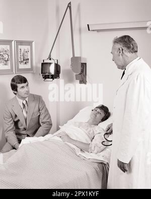1970S MÉDECIN DE L'HÔPITAL PARLANT À UNE PATIENTE COUCHÉE DANS LE LIT DE L'HÔPITAL ET SON MARI ASSIS AU CHEVET - M9133 HAR001 HARS CONJOINT ÉPOUX HYGIÈNE MALADIE COPY SPACE DEMI-LONGUEUR FEMMES PERSONNES QUI S'OCCUPENT DES HOMMES MALADIE PARTENAIRE B&W D'ÂGE MOYEN HOMME MOYEN PROFESSION DE SANTÉ FOURNISSEURS DE SERVICES DE PRÉVENTION DE LA SOUFFRANCE FOURNISSEURS DE SOINS PROFESSIONNELS DE GUÉRISON ET DE DIAGNOSTIC MÉDECINS PROFESSIONNELS DE LA SANTÉ DÉFICIENCE TRAITEMENTS GUÉRISSEURS DE CHEVET HÔPITAUX CONCEPTUELS CENTRE DE SOUTIEN DES MÉDECINS PRATICIENS MAUVAISE SANTÉ MALADE ADULTE MOYEN-ADULTE HOMME ADULTE MOYEN-ADULTE FEMMES PROFESSIONNELLES Banque D'Images
