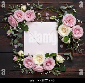 Enveloppe carrée entre roses rose et crème sur bois brun vue du dessus, maquette de mariage. Scène romantique avec enveloppe et fleurs pastel. Valent Banque D'Images