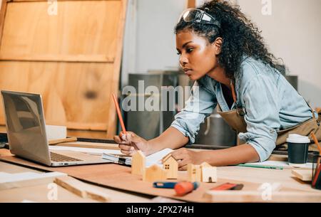 Carpenter america noir femme cheveux curly dessins sur papier travail avec ordinateur portable, jeune femme travaillant croquis emploi et l'apprentissage en ligne à Woodshop, National Banque D'Images