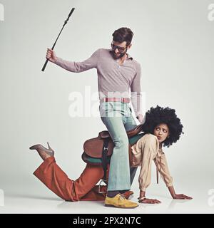 HES en charge maintenant. Photo en studio d'un homme attrayant en 70s, portant une jeune femme portant une selle tout en utilisant une récolte d'équitation. Banque D'Images