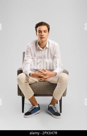 Jeune pensif beau homme en pantalon beige et chemise blanche assis dans un fauteuil en studio, il a ses doigts entremêtés et regardant droit vers l'avant. Banque D'Images