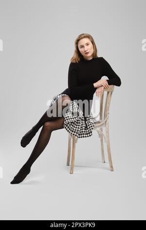 Femme pieds nus en col roulé et petite jupe assise sur une chaise en studio sur fond gris Banque D'Images