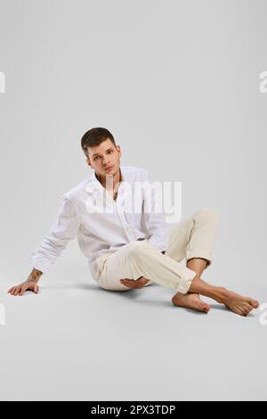 Portrait en studio complet d'un jeune homme pieds nus en chemise blanche et pantalon ivoire assis au sol Banque D'Images