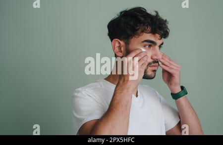 Vue latérale d'un homme barbu qui fait la routine matinale en utilisant un tampon de coton, en regardant loin se tenant isolé sur fond gris. Soins de la peau, toilettage Banque D'Images