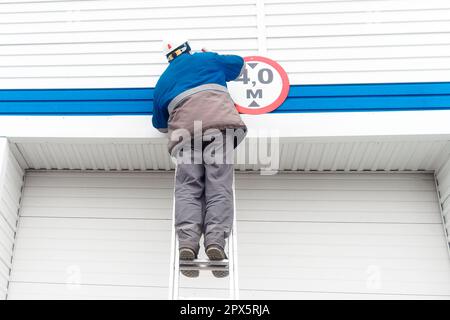 Un électricien mâle se tient sur une échelle et remplace une ampoule dans une lampe de rue sur la porte d'un bâtiment industriel ou d'un garage. Flux de travail authentique. Vue de l'arrière de bas en haut. Banque D'Images
