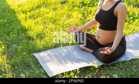 Une jeune femme enceinte caucasienne mignonne médite en s'asseyant sur un tapis sur la pelouse lors d'une soirée estivale ensoleillée. Concept de pacification Banque D'Images