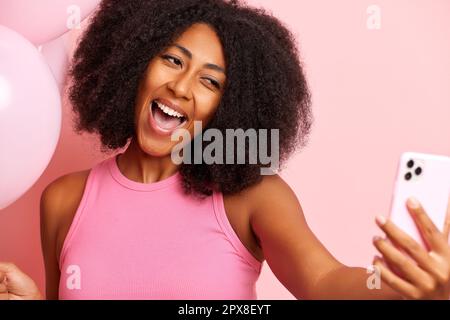 Charmante femme à la peau sombre tient un smartphone et fait un selfie, sourires heureux et largement, isolé à côté du mur rose. Photo de haute qualité Banque D'Images