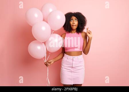 Jolie femme de race mixte, fait un fête isolé, tient des ballons, a croisé ses doigts dans un souhait et regarde, à l'espace de copie. Phot de haute qualité Banque D'Images