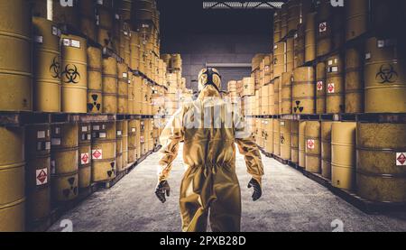 Homme en costume de protection jaune et masque à gaz, entrepôt plein de barils de métal jaune avec des symboles de danger. Banque D'Images