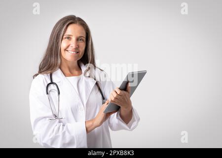 Femme médecin en bonne santé, sous le manteau, avec une tablette souriante à la caméra, posant sur fond gris studio, espace de copie Banque D'Images