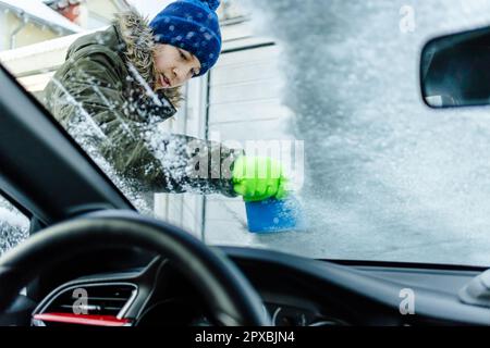 L'adolescent nettoie la voiture après une chute de neige, en enlevant la neige et en raclant la glace Banque D'Images