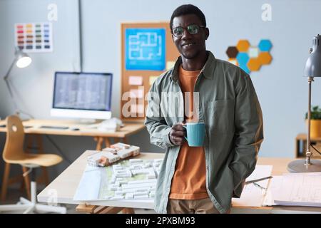 Portrait d'un architecte afro-américain qui boit du café tout en se tenant au bureau et en souriant à l'appareil photo Banque D'Images