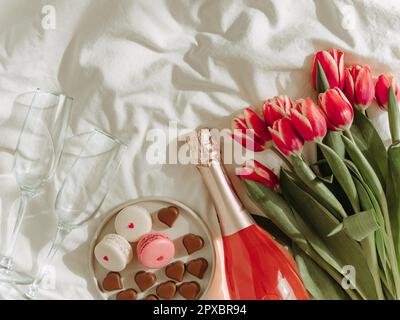 Bouquet de tulipes rouges, vin rose, verres à champagne, macarons et bonbons en forme de coeur sur draps blancs. Concept de la Saint-Valentin, date, mars 8. Ressort Banque D'Images