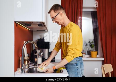 Homme d'âge moyen dans les lunettes laver les plats dans la cuisine le soir Banque D'Images