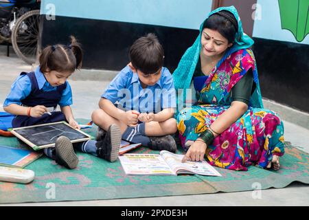 Un jeune enseignant indien aide les jeunes garçons et les jeunes filles à lire des livres et à écrire sur un tableau d'ardoise tout en étant assis sur le sol. Enfants portant une uniforme scolaire Banque D'Images