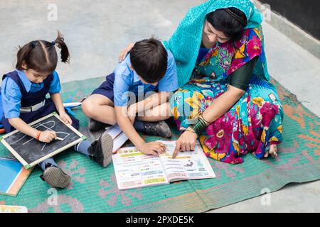 Un jeune enseignant indien aide les jeunes garçons et les jeunes filles à lire des livres et à écrire sur un tableau d'ardoise tout en étant assis sur le sol. Enfants portant une uniforme scolaire Banque D'Images