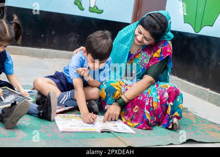 Un jeune enseignant indien aide un petit garçon étudiant avec un livre de lecture tout en étant assis sur le sol. Enfant portant l'uniforme scolaire étudiant à l'école gouvernementale. Banque D'Images