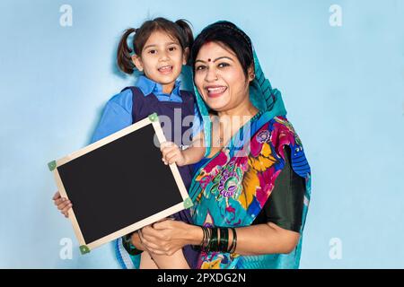 Jeune mère indienne heureuse avec sa petite fille mignonne portant l'uniforme de l'école et tenant un tableau d'ardoise vierge. Inde rurale, concept d'éducation. Banque D'Images