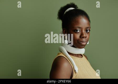 Jeune afro-américaine jolie femme avec un petit pain et un serpent enlaçant son cou regardant l'appareil photo tout en se tenant debout sur fond vert Banque D'Images