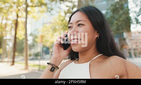 Jolie petite fille brune, vêtue d'un haut blanc et d'un Jean, assise sur un banc sur fond de paysage urbain, parlant sur un smartphone Banque D'Images