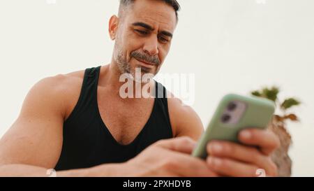 Un homme musclé d'âge moyen dans les vêtements de sport utilise un smartphone tout en étant assis sur la promenade sur fond de mer Banque D'Images