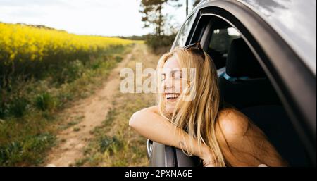 Jeune femme souriante assise en voiture et assise sur la fenêtre pendant son voyage en voiture et appréciant la belle journée de printemps dans la nature Banque D'Images