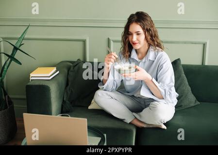 Week-end de détente à la maison: Jeune femme dans des vêtements confortables de la série de télévision, de profiter de temps libre avec un ordinateur portable et des collations sur le canapé. Un jeune Banque D'Images