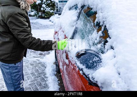 L'adolescent nettoie la voiture après une chute de neige, en enlevant la neige et en raclant la glace Banque D'Images