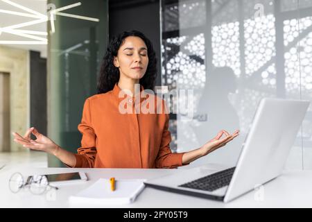 Jeune beau développeur web hispanique travaillant à l'intérieur d'un immeuble de bureau, femme se reposant méditant sur le lieu de travail avec les yeux fermés, femme employée utilisant un ordinateur portable pour écrire du code. Banque D'Images