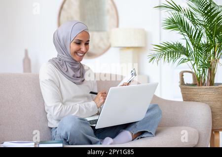 Jeune femme musulmane dans hijab freelance travail à partir de la maison en ligne. Assis sur le canapé en position lotus et tenant un ordinateur portable sur ses genoux. Il écrit des documents dans un dossier. Sourires, zoom sur la réunion. Banque D'Images