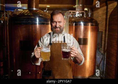 Brasseur senior satisfait posant pour caméra avec de la bière pâle et de la bière sombre pinte dans la main. Portrait de propriétaire de brasserie réussie annonçant craf fraîchement fait Banque D'Images