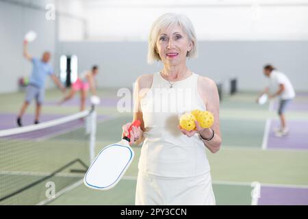 Femme âgée sportive souriante debout sur un terrain de pickleball intérieur Banque D'Images