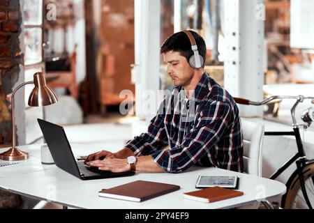 La musique me met toujours dans une humeur productive. un beau jeune homme d'affaires qui écoute de la musique tout en travaillant sur un ordinateur portable dans un bureau à l'intérieur de son atelier. Banque D'Images