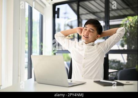 Un homme d'affaires asiatique millénaire fatigué s'assoit à son bureau, étirant ses bras pour se détendre après avoir terminé son travail. Banque D'Images